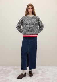 Suéter de punto con patrón de zigzag en blanco y negro, dobladillo acanalado rojo; combinado con una falda larga de mezclilla con abertura frontal y zapatos marrones.