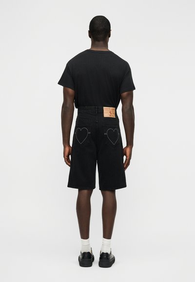 Μαύρα τζιν σορτς με τσέπες στην πλάτη σε σχήμα καρδιάς, συνδυασμένα με μαύρο μπλουζάκι και λευκές κάλτσες, φορεμένα με μαύρα παπούτσια.