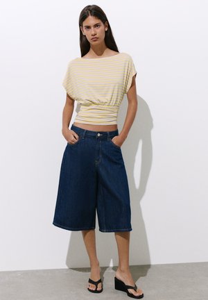 Femme debout, les mains dans les poches, portant un haut à manches courtes rayé jaune, un short en jean ample et foncé, et des sandales compensées noires.