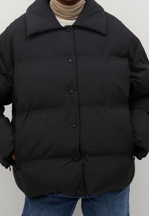 Doudoune noire avec boutons-pression, col large, portée sur un pull à col roulé beige et un jean bleu, mains dans les poches.