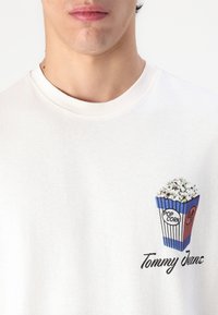 Hvit bomullsgenser med grafikk av en blå og rød popcornboks. "Tommy Jeans" er trykket i svart under designet.