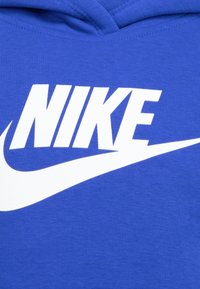 Modra kapuca z velikim belim Nike logotipom in žigom. Narejena iz teksturirane tkanine, ima kapuco in rebrasto obdelavo.