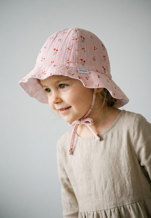 Junges Mädchen trägt einen rosa Sonnenhut mit Kirschenmuster und ein beiges Langarmkleid, lächelt leicht und schaut nach rechts.