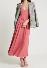 Lång rosa klänning med v-ringning och flödande kjol, parat med en grå rutig blazer. Modellen har på sig beige klackar. Slät tyg, lagerdesign.