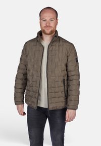 Giacca puffer di colore verde oliva con design trapuntato, chiusura con zip e tasche laterali. Indossata sopra una camicia di colore chiaro.