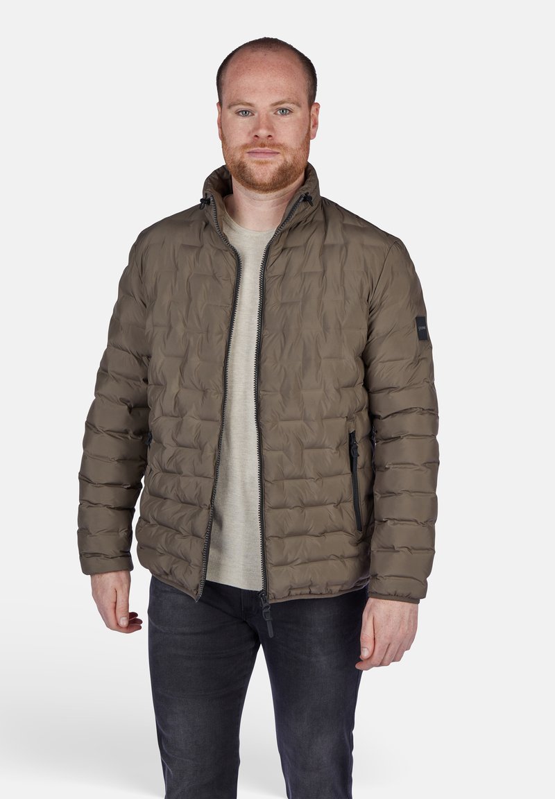 Giacca puffer di colore verde oliva con design trapuntato, chiusura con zip e tasche laterali. Indossata sopra una camicia di colore chiaro.