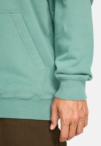 Mint green hoodie made of soft fabric with ribbed cuffs and hem, featuring a kangaroo pocket and a relaxed fit. Hand resting on the side.