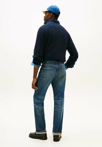 Pullover blu navy con finitura testurizzata, polsini in denim azzurro chiaro, jeans blu sbiaditi con dettagli di cucitura e scarpe nere.