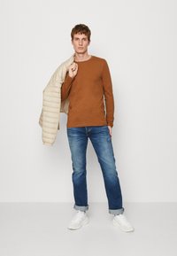Brun langermet skjorte, lys beige quiltet jakke over skuldrene, blå jeans og hvite sneakers med oppbrett.