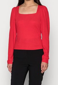 Röd långärmad topp med fyrkantig halsringning. Strukturerad tyg med en figurnära design. Kombination med svarta byxor som visar en kontrasterande look.