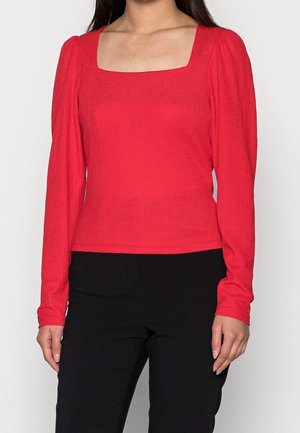 Top rosso a maniche lunghe con scollatura quadrata. Tessuto testurizzato con un design aderente. Abbinato a pantaloni neri, creando un contrasto visivo.