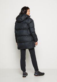 Moteris stovinti šonu, vilkinti juodą pūkinę striukę su gobtuvu, juodas kelnes ir juodus "Columbia" sportbačius ant baltos fono.