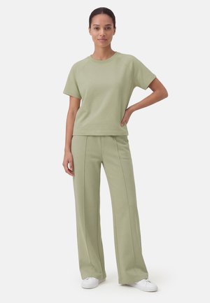 Nainen seisoo käsi lanteilla, yllään vaaleanvihreä lyhythihainen paita, vastaavat leveälahkeiset housut ja valkoiset tennarit valkoisella taustalla.