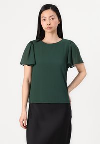 Grüne Chiffon-Bluse mit kurzen, gerafften Ärmeln, rundem Ausschnitt und lockerer Passform, kombiniert mit einem schwarzen Rock. Glattes, leichtes Material.