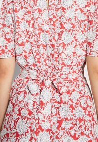 Korte mouwen jurk van rode stof met een wit bloemenpatroon, met een getailleerde taille en een strikdetail op de rug. Katoenmix materiaal.