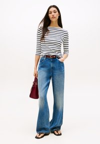 Ilgos mėlynos džinsinės kelnės su laisvu siluetu, derintos su juodai baltos spalvos dryžuota ilgomis rankovėmis palaidine ir maža burgundiško raudonio rankine.