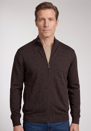 Ein Mann trägt einen dunkelbraunen Reißverschluss-Pullover über einem beigen Hemd, steht vor einem schlichten hellen Hintergrund und schaut geradeaus.