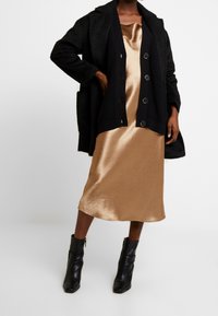 Black textured coat over a bronze satin dress, featuring a relaxed silhouette, button details, and black ankle boots with a sleek finish.