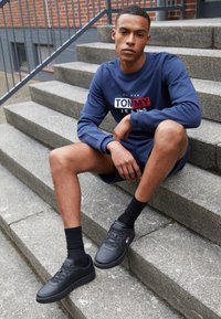 Sneakers neri con texture perforata, indossati con calzini neri, abbinati a una felpa blu con un design logo rosso e bianco.