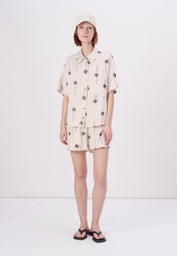 Chemise beige clair à manches courtes et shorts assortis avec imprimé de palmiers noirs ; comprend une casquette blanche texturée et des tongs noires.