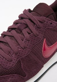 Nahaufnahme eines dunkelvioletten Nike-Sneakers mit strukturiertem Stoff, geflochtenen Schnürsenkeln, perforiertem Wildleder und einem glänzenden roten Nike-Swoosh-Logo.