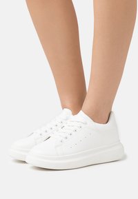 Hvide sneakers med en glat læderoverflade, rund tå og snørebåndsdesign; har perforerede detaljer på siderne og en tyk sål.