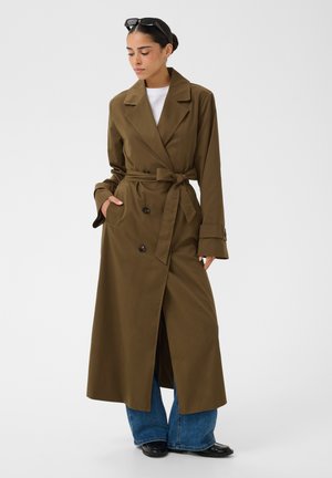 Femme portant un long trench-coat marron ceinturé, jean bleu à jambes larges, chaussures noires, chemise blanche et lunettes de soleil sur la tête.