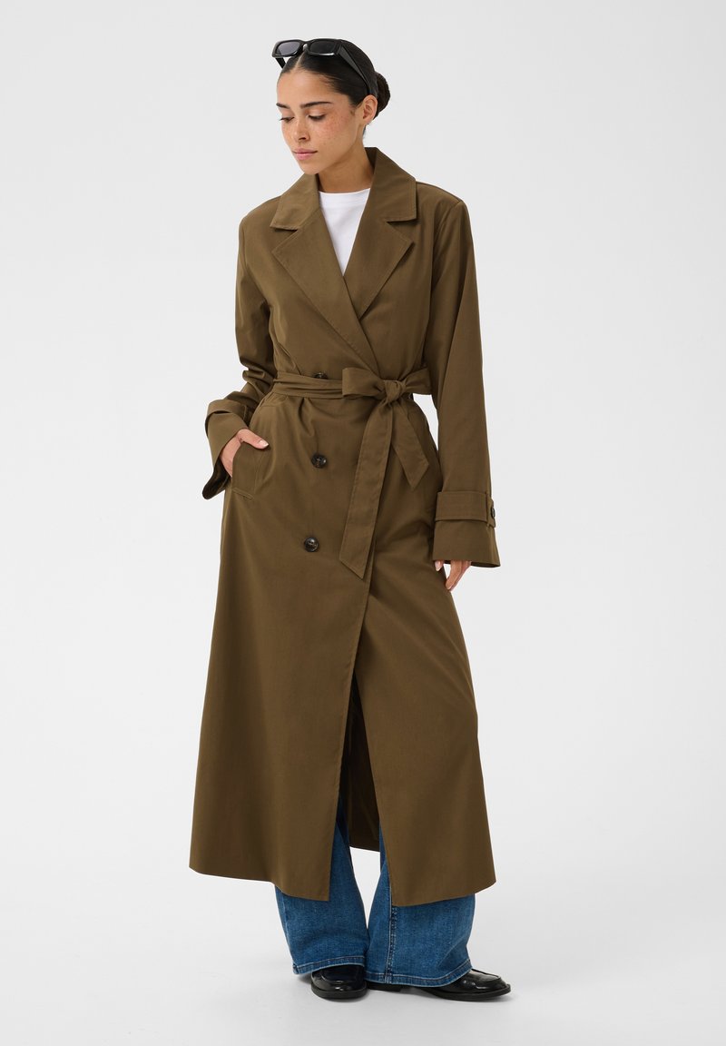 Femme portant un long trench-coat marron ceinturé, jean bleu à jambes larges, chaussures noires, chemise blanche et lunettes de soleil sur la tête.