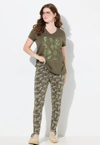 T-shirt verde oliva con scollo a V e stampa botanica in verde chiaro; abbinato a pantaloni elasticizzati floreali verde oliva e scarpe beige con lacci.