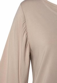 Beige langwerpige top met lange mouwen, gemaakt van zachte stof, met geplooide details op de schouder en een ronde halslijn. Gladde textuur en relaxte pasvorm.