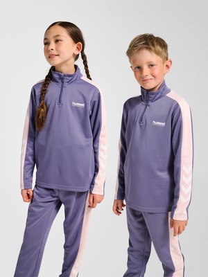 To børn iført matchende lilla træningsdragter med lyserøde striber, stående og vendt mod kameraet foran en ensfarvet baggrund.