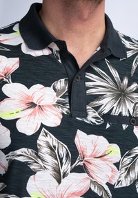 Polo de manga corta con un fondo oscuro y grandes patrones florales en rosa y blanco, con cuello abotonado y un bolsillo en el pecho.