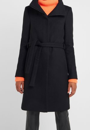 Manteau en laine noir ceinturé porté sur un pull à col roulé côtelé orange vif et un pantalon noir, présenté sur une personne debout.