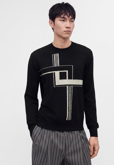 Jeune homme portant un pull noir avec un motif géométrique blanc et une clé grecque, associé à un pantalon rayé vertical gris et noir.