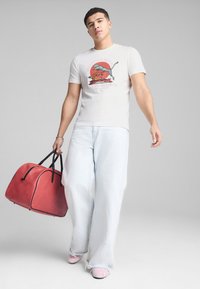 Una camiseta en gris claro con un estampado gráfico colorido, combinada con vaqueros de tiro ancho en azul claro y un gran bolso de viaje de cuero rojo con acentos negros.