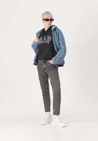 Blå fleecejakke over en svart hettegenser med "GAP"-logo, grå jeans og hvite sneakers. Glatt tekstur, avslappet passform og uformell stil.