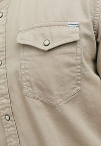 Beige overhemd met knopen, voorzien van een borstzak met drukknoopsluiting, zichtbare stiksels en een merklabel bij de zak. Zachte, gestructureerde stof.