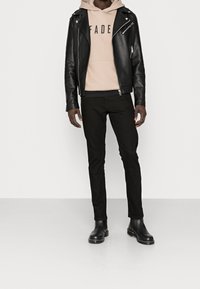 Giacca da biker in pelle nera sopra un hoodie beige con il testo "FADE", abbinata a pantaloni slim-fit neri e stivali Chelsea neri.