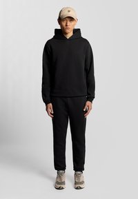 Sort hættetrøje og matchende sweatpants, båret med en beige cap med et logo, samt beige og grønne sneakers. Glat tekstur.