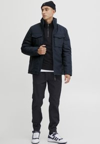 Marineblaue Steppjacke mit zwei Fronttaschen und hohem Kragen, getragen über einem schwarzen Reißverschlussoberteil, kombiniert mit schwarzen Hosen und weißen Sneakers.