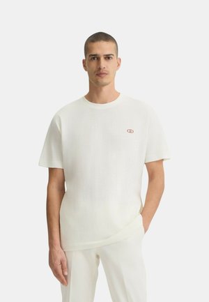 T-shirt bianco a maniche corte realizzato in tessuto morbido, con scollatura rotonda e piccolo logo rosso sul petto, abbinato a pantaloni di colore chiaro.
