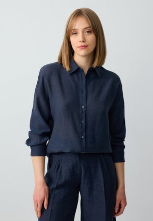 Femme jeune avec des cheveux châtains clairs à la longueur des épaules, portant une chemise à manches longues bleu marine à boutons et un pantalon assorti, sur un fond uni.