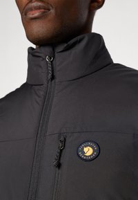 Black zip-up jacket with a high collar, featuring a gold and navy logo patch on the chest and side zipper pocket. Smooth texture.