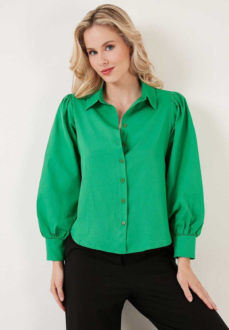 Camicia verde con bottoni e maniche a sbuffo, realizzata in un tessuto liscio. Presenta un colletto e bottoni rivestiti sul davanti. Indossata con pantaloni neri.