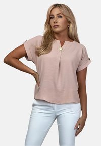 Jasnoróżowa bluzka z krótkim rękawem z dekoltem w kształcie litery V, teksturowana tkanina i ozdobny złoty guzik. Połączona z białymi spodniami.