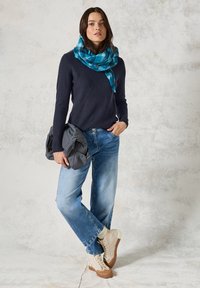 Pull en maille bleu marine avec motif diagonal texturé, jean bleu clair, baskets beiges et écharpe multicolore bleue. Tenant une veste grise.
