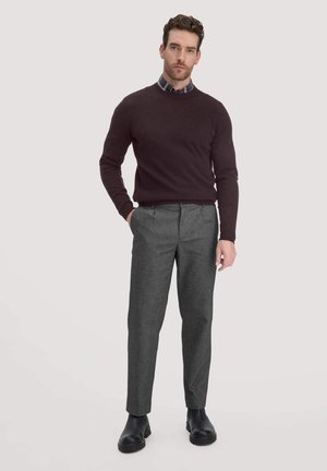 Dunkelmaroones Herrenpullover mit Rundhalsausschnitt, kombiniert mit grauen, maßgeschneiderten Hosen. Schwarze Knöchelstiefel ergänzen das Outfit. Ein karierter Hemd ist darunter sichtbar.