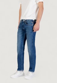 Jeans in denim blu con vestibilità dritta, cinque tasche e cuciture a contrasto, abbinati a una t-shirt bianca e sneakers bianche.