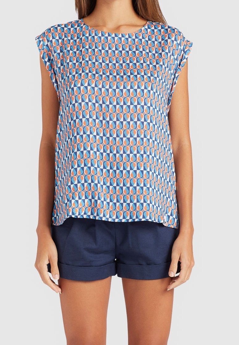 Femme portant un haut à manches courtes à motifs avec des formes bleues, orange et blanches et un short bleu marine à revers sur un fond uni.