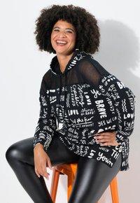 Sudadera negra con patrones de texto blanco, con una malla en las mangas, combinada con leggings negros brillantes. Modelo sentada en un taburete naranja.